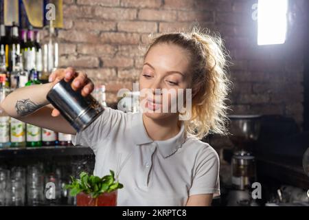 Junge, wunderschöne Barkeeperin streut den Cocktail mit Puderzucker. Die genaue Einhaltung des Rezepts bei der Zubereitung von Cocktails ist der Schlüssel zu s Stockfoto