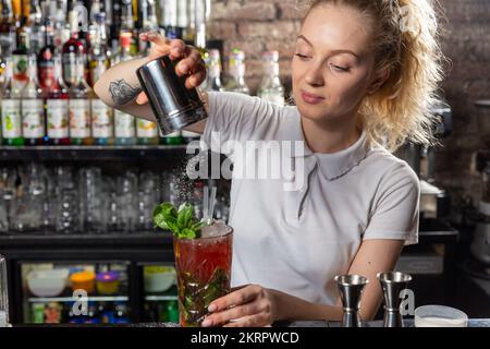 Junge, wunderschöne Barkeeperin streut den Cocktail mit Puderzucker. Die genaue Einhaltung des Rezepts bei der Zubereitung von Cocktails ist der Schlüssel zu s Stockfoto