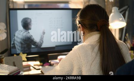 Ein junges asiatisches Mädchen sitzt am Tisch in einem gemütlichen Zimmer und lernt neues Material. Das Schulmädchen hört sich eine Online-Mathematik- und Studienstunde auf dem pc an. Der erfahrene Lehrer spricht mit dem Schüler und zeigt Gleichungen an Bord Stockfoto