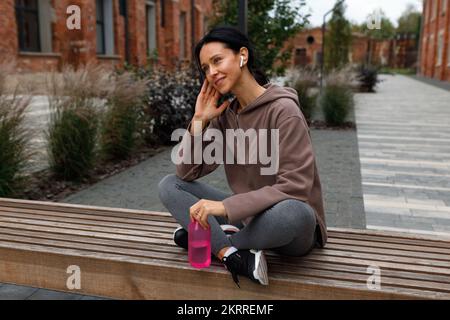 Wunderschöne Fitnessfrau mit einer Sportflasche Wasser in den Händen, die motivierende Musik in den Kopfhörern hört, während sie nach dem Workout im Freien auf der Bank sitzt Stockfoto