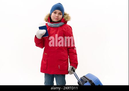 Glücklicher Reisedanker in rotem Parka, Jeans, Wollhandschuhen, macht Winterferien, geht mit Flugticket und blauem Koffer spazieren Stockfoto