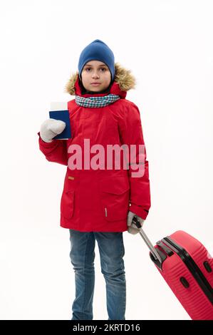 Ein junger Reisender mit rotem Parka und blauer Jeans läuft mit einer Bordkarte und einem Koffer auf weißem Hintergrund Stockfoto