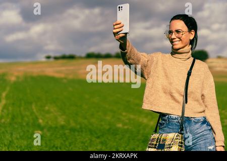 Ein junges brünettes Mädchen mit Brille und eine schöne Tasche, die an ihrer Schulter hängt, lächelt, während sie ihr Smartphone mit der Hand hält, um ein Selfie zu machen Stockfoto