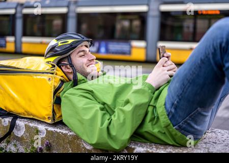 Junger Mann, der auf einer niedrigen Mauer in der Stadt ruht, schneller Transport in der Stadt von Essen, Getränken und Dokumenten Stockfoto