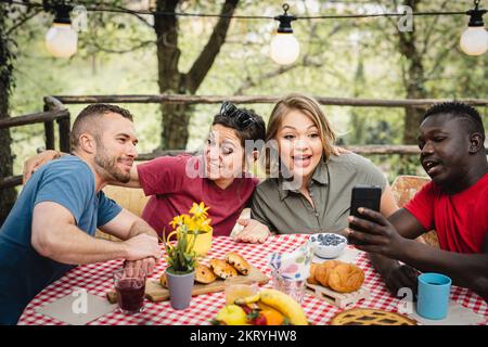 Glückliche Freunde frühstücken im Hinterhof, junger Influencer, der sein Smartphone benutzt, um Inhalte in sozialen Medien mit Fotos von Essen zu erstellen, multiethnische c Stockfoto