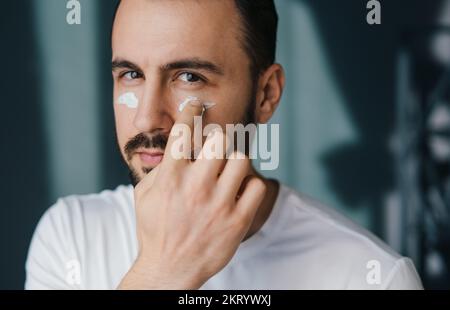 Attraktiver junger Mann, der zu Hause nährende Creme aufträgt und in die Kamera schaut. Genießen Sie die Morgenroutine. Schönheitsbehandlung. Dermatologie Stockfoto