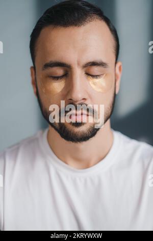 Nahaufnahme des Frontsichtbildes eines jungen bärtigen Mannes mit aufgetragener Augenklappe auf grauem Hintergrund. Gesichtsbehandlung, Kosmetik, Schönheitskonzept Stockfoto