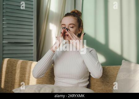 Porträt einer jungen Weißen mit Ingwerhaaren und frischer Sommersprossen, die auf dem Sofa sitzt und das Gesicht berührt. Fauler Morgen. Gemütliche Wohnung. Haut Stockfoto