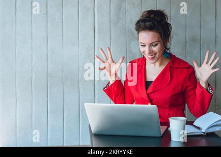 Eine Erwachsene, weiße Führungskraft, die mit erhobenen und offenen Händen glücklich reagiert und lächelt, während sie an ihrem Laptop arbeitet und in einem Café sitzt. Stockfoto