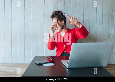 Weiße Erwachsene Frau, in einem roten Blazer mit ihrem Laptop, die in einer Cafeteria sitzt, sich nach hinten lehnt und ihre Überraschung ausdrückt, die ihr Gesicht mit den Händen bedeckt, Stockfoto