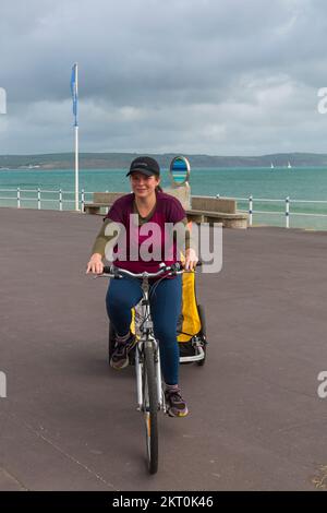 Junge Frau, die im Oktober auf dem Fahrrad entlang der Promenadenpromenade in Weymouth, Dorset, UK fährt Stockfoto