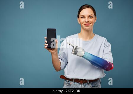 Glückliche junge Frau mit Armprothese, die auf den Smartphone-Bildschirm zeigt und eine neue App empfiehlt Stockfoto