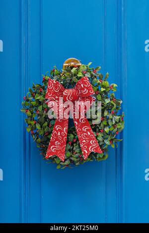 Traditioneller grüner und roter Weihnachtskranz hängt an der blauen Eingangstür des Hauses. Traditionelle Einrichtung zu Weihnachten Stockfoto