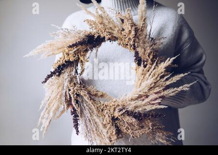 Stilvoller Herbstkranz. Hände mit trockenem Gras, Wildblumen und Weizenkranz Stockfoto