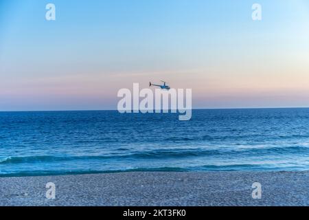 Ein Hubschrauber fliegt über das Meer in der Nähe eines Sandstrands Stockfoto