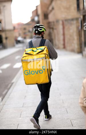 Spanien, Salamanca - 3. April 2022: Lieferung von Lebensmitteln und Lebensmitteln. Der Fahrradkurier mit gelbem Thermo-Rucksack, die Firma Glovo, liefert das nötige Stockfoto