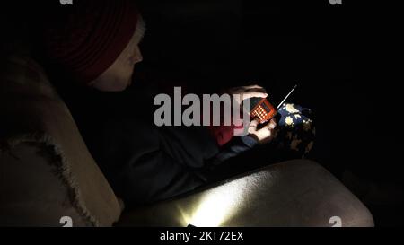 Ukraine: Großmutter hält während russischer Terroranschläge Radio in den Händen, während sie im Dunkeln des Blackouts sitzt. Seniorin in Winterkleidung im Dunkeln Stockfoto