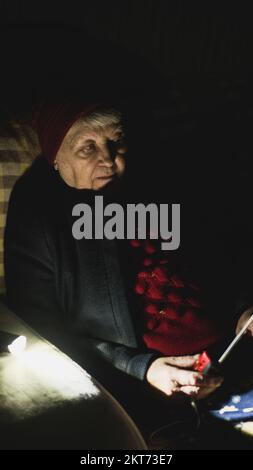 Ukraine: Großmutter hält während russischer Terroranschläge Radio in den Händen, während sie im Dunkeln des Blackouts sitzt. Seniorin in Winterkleidung im Dunkeln Stockfoto