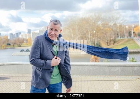 Porträt eines glücklichen Mannes mittleren Alters mit kurzen Haaren, der eine graue Jacke trägt, und einem fliegenden blauen Schal, der neben einer Betonattika steht. Stockfoto