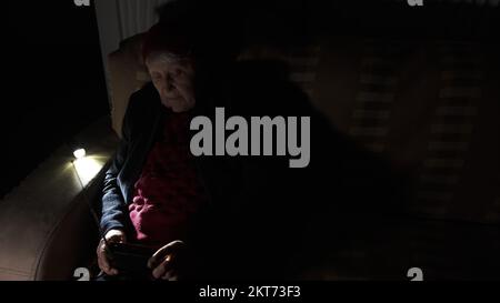 Ukraine: Großmutter hält während russischer Terroranschläge Radio in den Händen, während sie im Dunkeln des Blackouts sitzt. Seniorin in Winterkleidung im Dunkeln Stockfoto