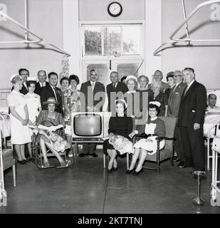 1964, Geschichte, Krankenschwestern und medizinisches Personal versammeln sich zu einem Gruppenfoto, um ein neues Fernsehgerät zu feiern, das auf einer Krankenhausstation in England, Großbritannien, eintrifft. Da Fernsehgeräte in dieser Zeit teuer waren, war es ein großes Ereignis für Krankenhäuser und Schulen, eines zu erhalten. Beachten Sie, dass ein eigenständiger Aschenbecher aus Edelstahl auf der rechten Seite wie in dieser Zeit zu sehen ist, da Rauchen immer noch zu bestimmten Zeiten auf vielen Krankenhausstationen erlaubt war. Stockfoto