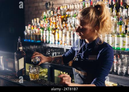 Junge, wunderschöne Barkeeperin gibt Limonade zu einem Cocktailglas. Limonade ist ein Teil vieler Cocktails. Stockfoto