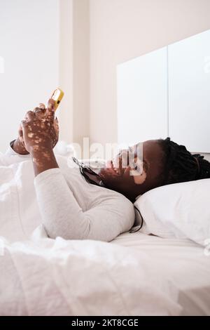Glückliche schwarze Frau mit weißer Pigmentierung, die im Schlafzimmer auf dem Handy surft Stockfoto