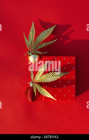 Goldene Cannabisblätter, Marihuana mit Weihnachtsdekor und Geschenkbox auf rotem Hintergrund. Das Konzept einer roten Feiertagskulisse für Plakate Stockfoto