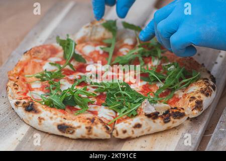 Hände mit Plastikhandschuhen Fügen Sie frische Raketenblätter auf eine Pizza mit Tomatensoße und Mozzarella auf einem Holzbrett hinzu, Nahaufnahme Stockfoto