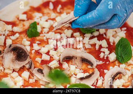 Hand mit Plastikhandschuh, um Pizza mit Tomatensoße, frischem Mozzarella, Pilzen und grünen Basilikumblättern auf einem Holzbrett zuzubereiten, Nahaufnahme Stockfoto