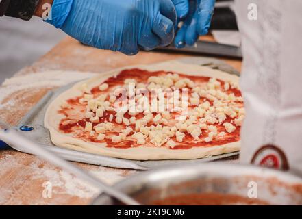 Hände mit Kunststoffhandschuhen Fügen Sie frischen Mozzarella auf eine Pizza mit Tomatensoße auf einem Holzbrett mit Zutaten hinzu, Nahaufnahme Stockfoto