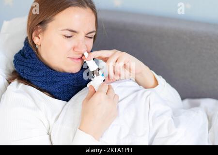 Junge Frau mit schwerer Erkältung, die im Bett unter der Decke liegt und Nasentropfen einnimmt Stockfoto