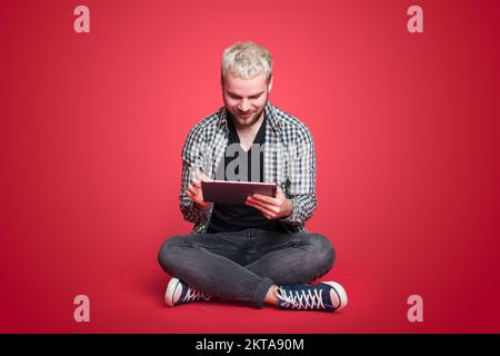 Porträt eines jungen Mannes, der auf dem Boden sitzt, mit gekreuzten Beinen und sich auf seinem Tablet etwas genau ansieht, isoliert vor rotem Hintergrund Stockfoto