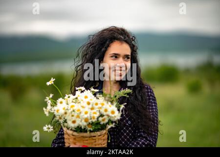 Eine Frau steht auf einem grünen Feld und hält einen Korb mit einem großen Strauß Gänseblümchen in den Händen. Im Hintergrund gibt es Berge und einen See. Stockfoto