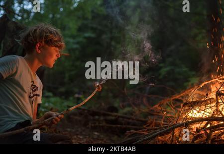 Ein junger, weißer Teenager, der abends die Fleischwurst über einem großen Lagerfeuer brät. Konzeptbild: Camping, Reisen und glückliche Teenager. Stockfoto