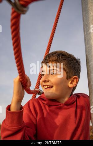 Porträt eines fröhlichen, lächelnden, süßen Jungen, der bei Sonnenuntergang an einem Seil hängt. Positives Konzept Stockfoto