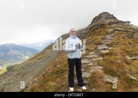 Eine grauhaarige Frau in Sportbekleidung steht auf einem Berg und schaut in die Kamera. Österreich, Alpen. Hochwertiges Foto Stockfoto