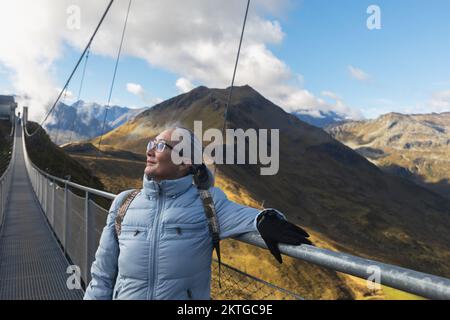 Eine Erwachsene grauhaarige Frau steht auf einer Hängebrücke hoch in den Bergen über einem Abgrund in den österreichischen Alpen. Hochwertiges Foto Stockfoto