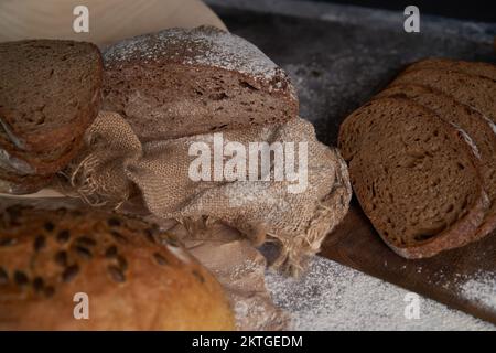 Draufsicht auf verschiedene Brotsorten, geschnitten und ganz, Mehl, zerknittertes Papier und Latte auf einem Holztisch. Großer Winkel. Stockfoto