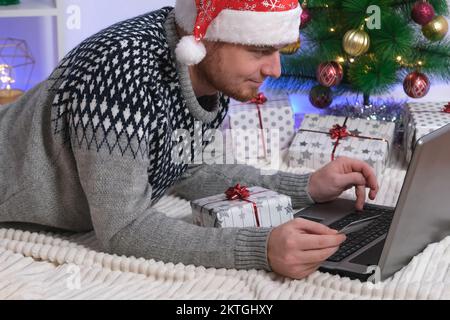 Mann mit Laptop und Kreditkarte kauft Geschenke für Neujahr 2023, Weihnachten. Ein Mann im Weihnachtsmütze auf dem Hintergrund von Weihnachtsbaum, Girlanden, Geschenken mit einem Laptop. Cybersicherheit vor Betrügern. Neon-Bokeh. Stockfoto