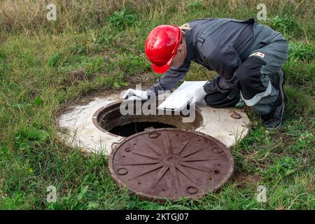 Ein Mann, der Klempner in Overalls arbeitet, die über einem Brunnen gebogen sind, fixiert die Messungen und die Messwerte des Wasserzählers. Stockfoto