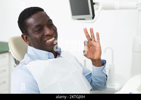 Ein lächelnder Afroamerikaner, der ein gutes Zeichen zeigt, während er in der Zahnarztpraxis sitzt Stockfoto