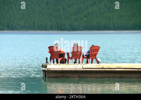 Banff, Alberta, Kanada - 2018. Juni: Drei Personen sitzen auf Stühlen auf einem Holzsteg, der auf dem Wasser des Lake Minnewanka schwimmt Stockfoto