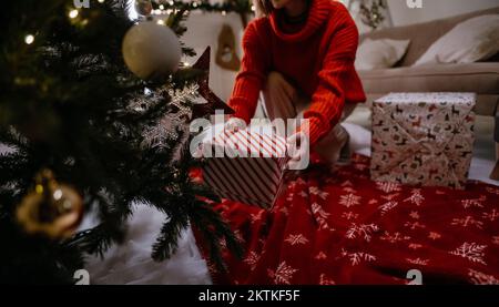 Nahaufnahme einer unbekannten Frau, die an einem gemütlichen Abend ein Geschenk unter dem Weihnachtsbaum hält Stockfoto