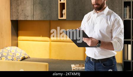 Ein bärtiger Einzelhändler macht sich Notizen, während er im Möbelgeschäft arbeitet. Schnittansicht des Vertriebsleiters, der auf einem Tablet schreibt, während er vor dem Schlafzimmermöbel steht. Konzept des Arbeitsplatzes. Stockfoto