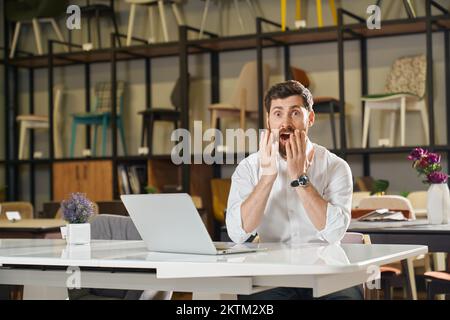 Der Manager in einem weißen Hemd war erstaunt, als er im Möbelgeschäft auf einem Laptop surfte. Porträt eines geschossenen männlichen Einzelhändlers, der aus Gewinnergebnissen oder Karrieremöglichkeiten heraus arbeitet, während er in Innenräumen arbeitet. Emotionsbegriff. Stockfoto