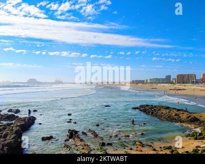 Menschen, die sich an sonnigen Tagen am Matosinhos Sea Beach entspannen, Stadtbild mit moderner Architektur und Hafen Leixoes im Hintergrund, Porto, Portugal Stockfoto