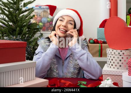 Sie spricht mit einem Mobiltelefon, lächelnde Frau bereitet Geschenke für Feiern in bunten Geschenkkartons auf ihrem Tisch vor Stockfoto