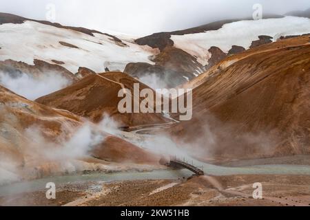 Brücke und dampfende Bäche zwischen farbenfrohen Rhyolitenbergen und Schneefeldern, geothermisches Gebiet Hveradalir, Kerlingarfjöll, isländische Highlands, Icel Stockfoto