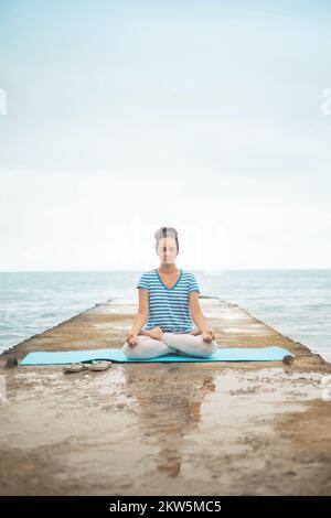 Frau Yoga-Lehrerin, die in Lotusstellung sitzt, zeigt sich mudra und bewundert die Küstenlandschaft Stockfoto